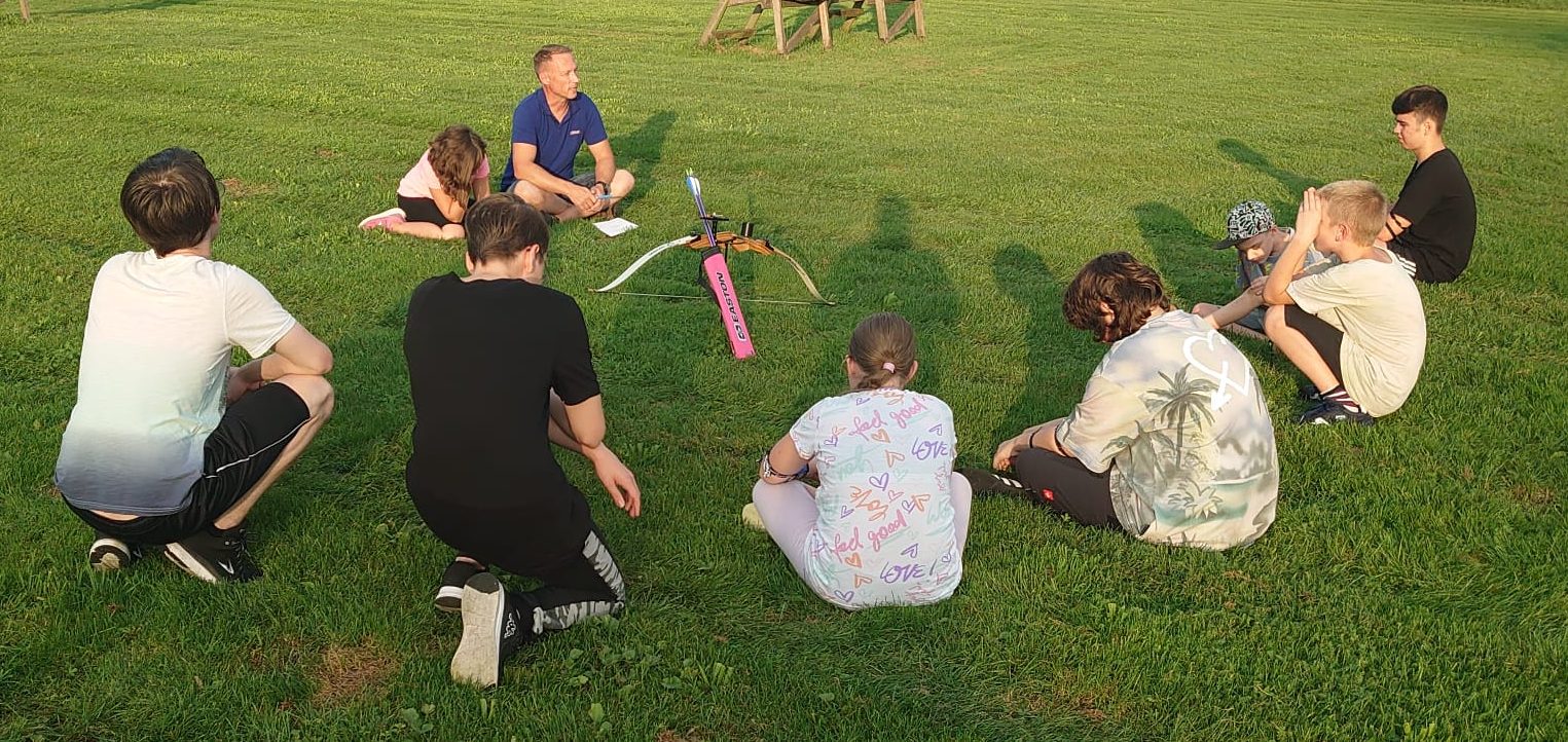 Sieben Menschen sitzen auf einer Bogensportwiese im Kreis und unterhalten sich.